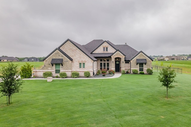 french country inspired facade featuring brick siding, fence, a front lawn, a shingled roof, and stone siding