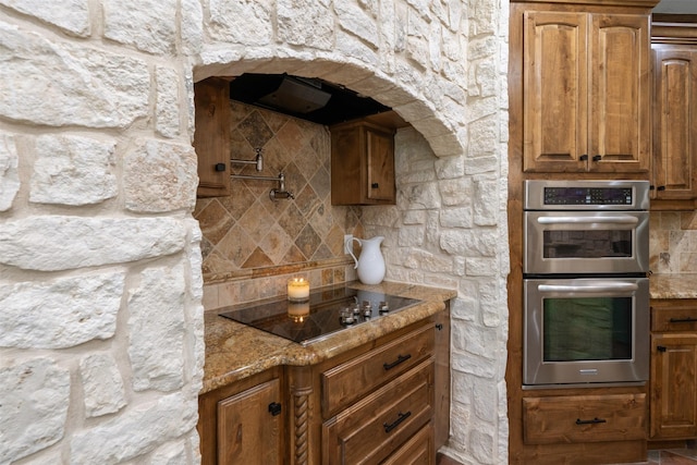 kitchen featuring brown cabinetry, double oven, light stone countertops, and black electric cooktop