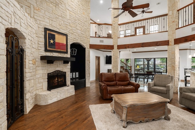 living room with a high ceiling, a stone fireplace, a ceiling fan, decorative columns, and wood finished floors
