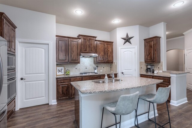 kitchen featuring appliances with stainless steel finishes, a sink, dark wood finished floors, under cabinet range hood, and a center island with sink
