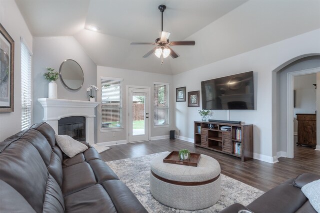living area featuring a glass covered fireplace, wood finished floors, baseboards, ceiling fan, and vaulted ceiling