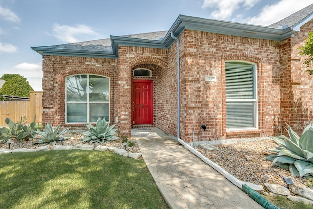 doorway to property with brick siding, roof with shingles, and fence