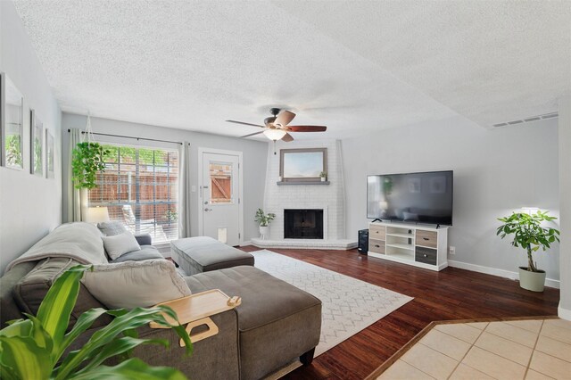 tiled living room with visible vents, a brick fireplace, baseboards, a textured ceiling, and a ceiling fan