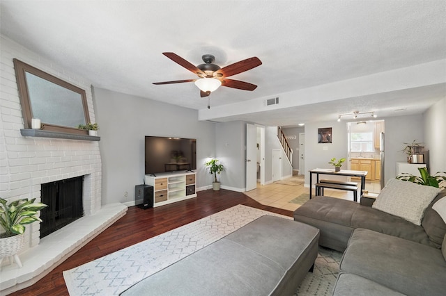 living area featuring ceiling fan, a brick fireplace, wood finished floors, visible vents, and stairway
