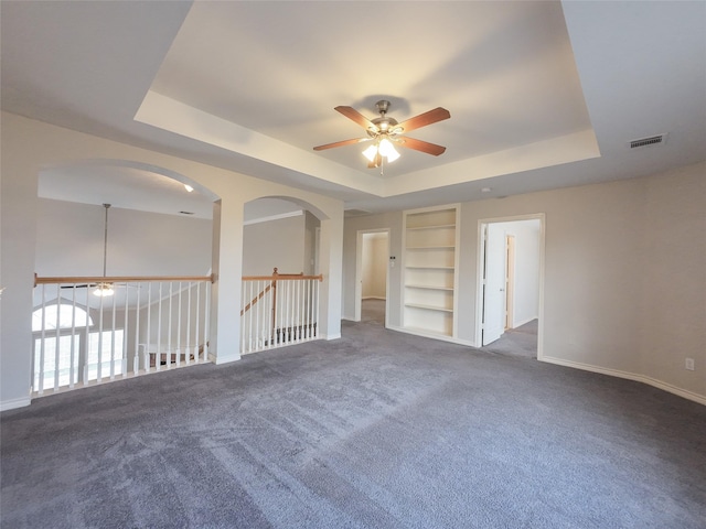carpeted empty room with visible vents, built in features, baseboards, a tray ceiling, and a ceiling fan
