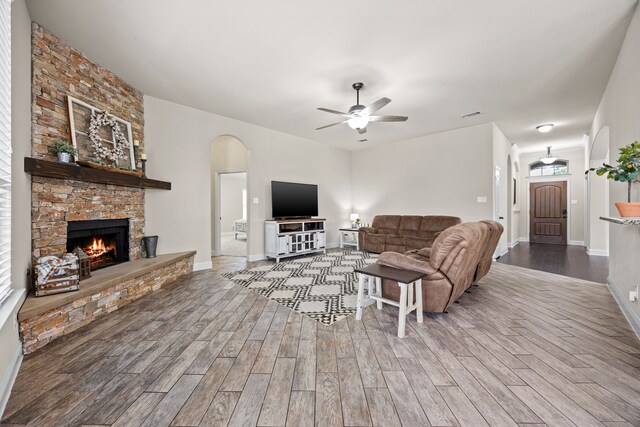 living room featuring wood finished floors, a stone fireplace, baseboards, ceiling fan, and arched walkways
