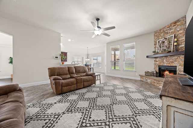 living room featuring baseboards, ceiling fan, a stone fireplace, and wood finished floors