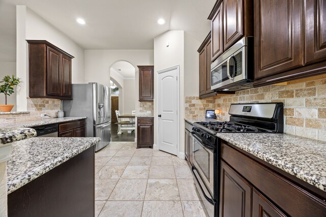 kitchen with dark brown cabinetry, light stone counters, arched walkways, backsplash, and stainless steel appliances