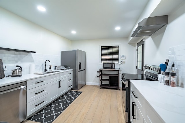 kitchen with light wood-type flooring, wall chimney exhaust hood, tasteful backsplash, a sink, and appliances with stainless steel finishes
