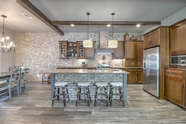 kitchen featuring stainless steel appliances, beam ceiling, brown cabinets, light stone countertops, and brick wall