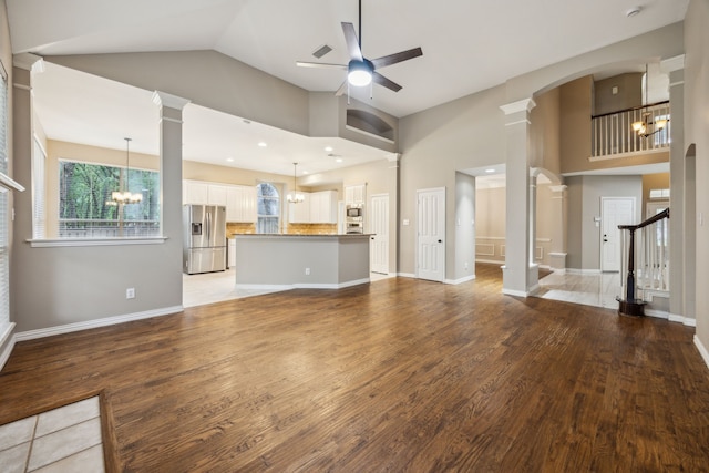 unfurnished living room featuring ornate columns, light wood finished floors, ceiling fan with notable chandelier, and high vaulted ceiling