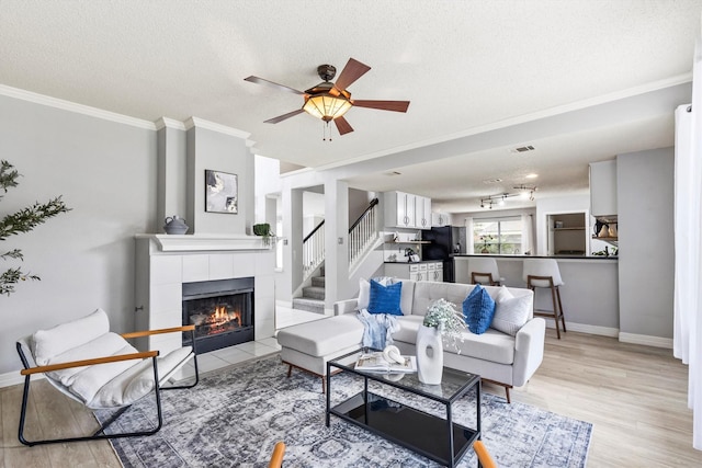 living area with ceiling fan, a tiled fireplace, ornamental molding, and light wood-type flooring