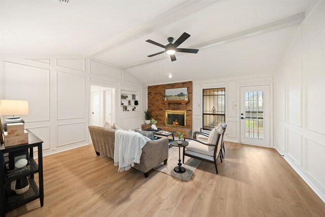 living area with ceiling fan, light wood-style floors, a brick fireplace, a decorative wall, and vaulted ceiling with beams