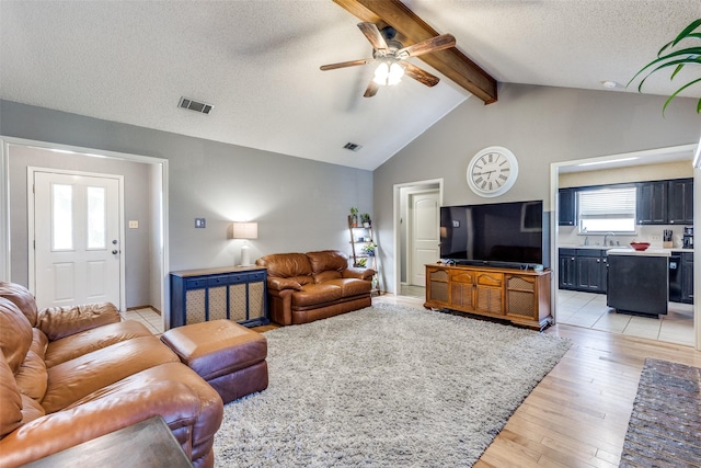 living area with ceiling fan, a textured ceiling, visible vents, lofted ceiling with beams, and light wood-style flooring