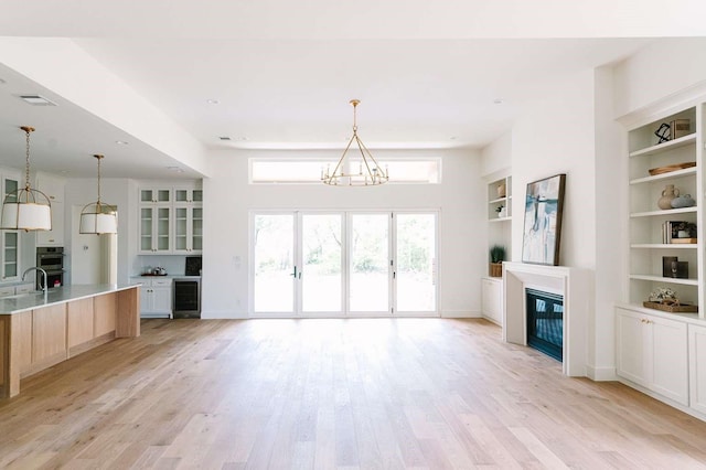 unfurnished living room featuring built in features, light wood-style flooring, a glass covered fireplace, and a chandelier
