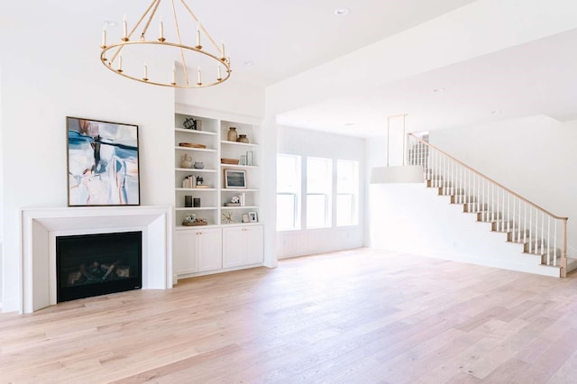 unfurnished living room with an inviting chandelier, a fireplace, built in shelves, light wood-type flooring, and stairway