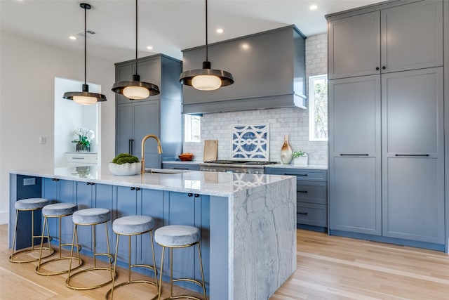 kitchen featuring backsplash, light stone countertops, a center island with sink, a sink, and light wood finished floors