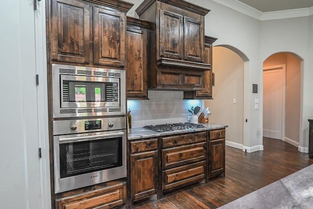 kitchen with decorative backsplash, appliances with stainless steel finishes, arched walkways, dark brown cabinets, and ornamental molding