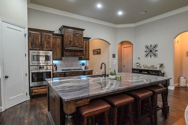 kitchen featuring a sink, a kitchen breakfast bar, a center island with sink, arched walkways, and stainless steel appliances