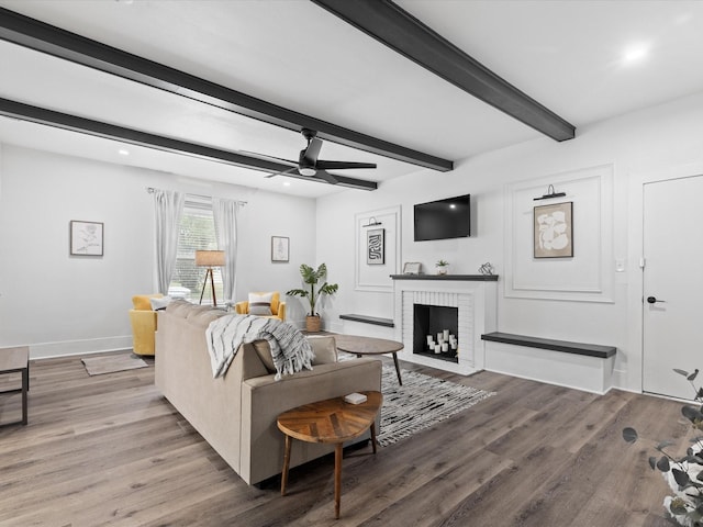 living area featuring a ceiling fan, a brick fireplace, wood finished floors, baseboards, and beamed ceiling