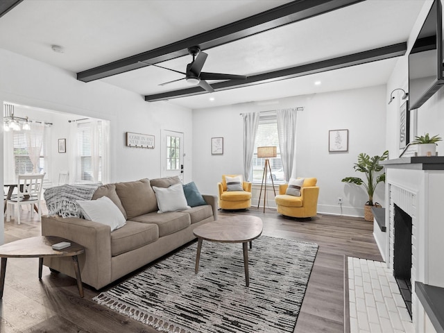 living room featuring wood finished floors, beamed ceiling, baseboards, a fireplace with flush hearth, and ceiling fan with notable chandelier