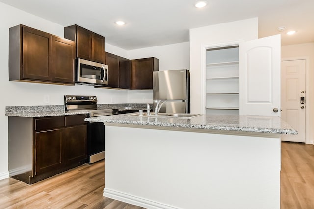kitchen featuring light stone countertops, a center island with sink, dark brown cabinets, light wood finished floors, and stainless steel appliances