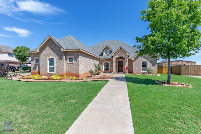 french country style house featuring brick siding, roof with shingles, fence, and a front yard