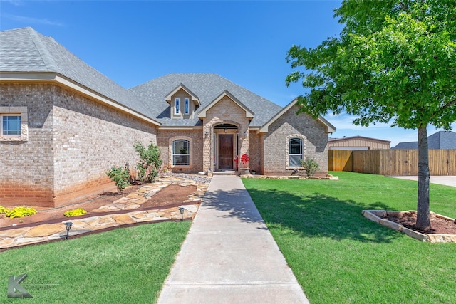 french country style house with brick siding, roof with shingles, fence, and a front yard