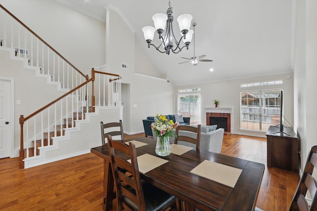 dining area featuring a fireplace, wood-type flooring, high vaulted ceiling, ceiling fan with notable chandelier, and stairs