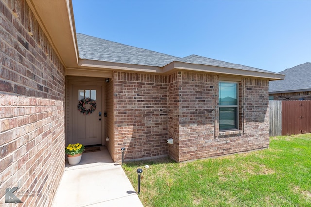 property entrance with brick siding, roof with shingles, fence, and a lawn