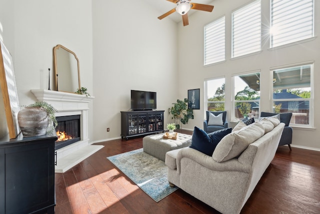 living area with baseboards, a towering ceiling, ceiling fan, a glass covered fireplace, and dark wood-style floors