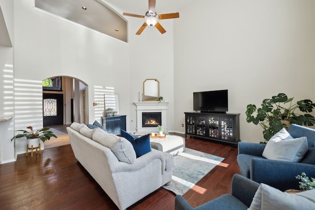 living area with hardwood / wood-style floors, ceiling fan, arched walkways, a high ceiling, and a glass covered fireplace