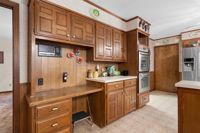 kitchen featuring brown cabinetry, crown molding, light countertops, built in desk, and appliances with stainless steel finishes