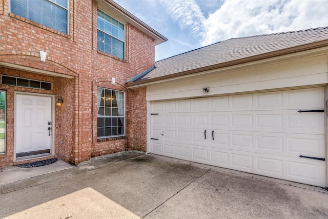 entrance to property featuring a shingled roof, concrete driveway, and brick siding