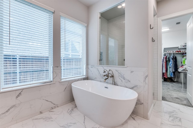 full bath with a walk in closet, marble finish floor, a wainscoted wall, a freestanding tub, and tile walls