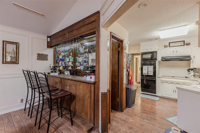 bar featuring a dry bar, light wood-style floors, a textured ceiling, under cabinet range hood, and black appliances
