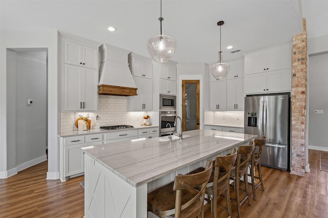 kitchen with a kitchen island with sink, wood finished floors, premium range hood, baseboards, and stainless steel appliances