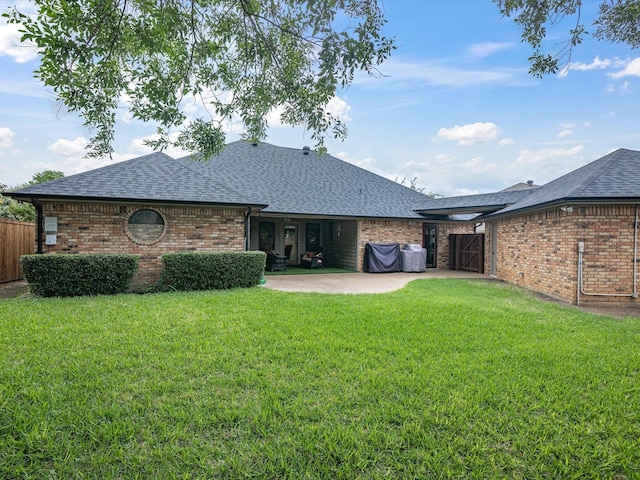 back of house with brick siding, roof with shingles, a lawn, and a patio