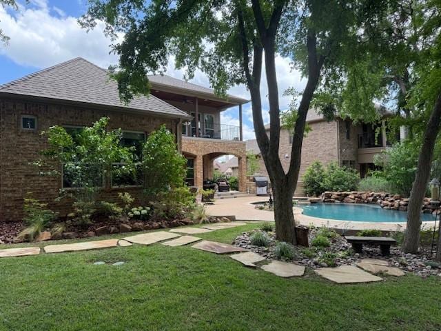 back of property with a shingled roof, brick siding, an outdoor pool, a balcony, and a lawn