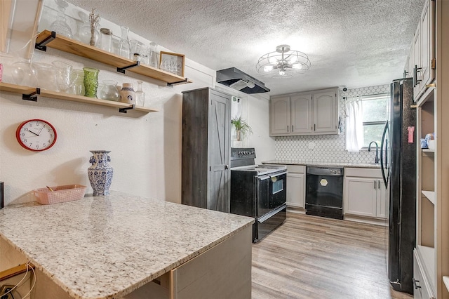 kitchen featuring wall chimney range hood, light wood-style floors, black appliances, a peninsula, and an inviting chandelier