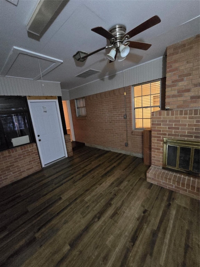 unfurnished living room with ceiling fan, attic access, brick wall, a brick fireplace, and wood finished floors