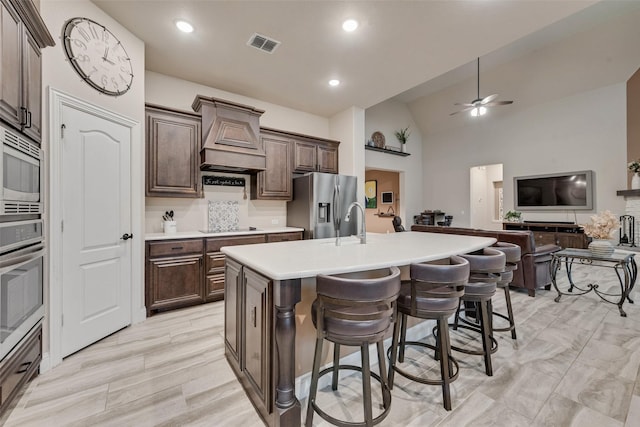 kitchen with stainless steel appliances, a kitchen island with sink, a breakfast bar area, a ceiling fan, and premium range hood
