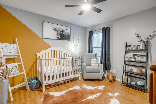 bedroom with a ceiling fan, a nursery area, wood finished floors, baseboards, and a textured wall