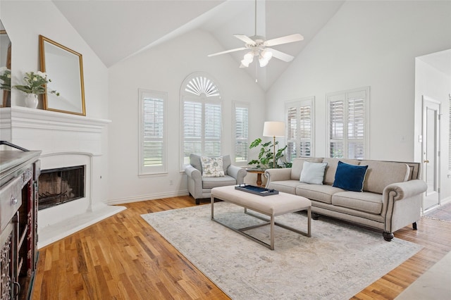 living room featuring a ceiling fan, high vaulted ceiling, baseboards, a fireplace with raised hearth, and light wood-style flooring