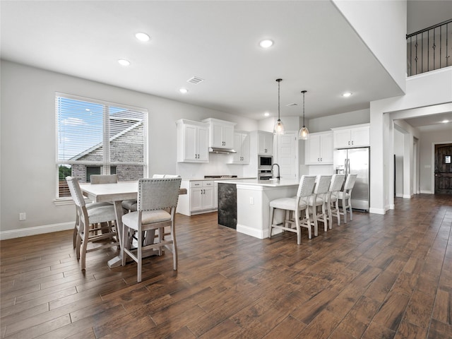 kitchen featuring dark wood-style flooring, appliances with stainless steel finishes, a breakfast bar area, under cabinet range hood, and a center island with sink