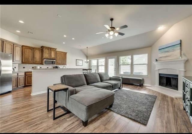 living room with a ceiling fan, vaulted ceiling, a fireplace with raised hearth, and light wood-style flooring