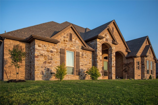 french country style house featuring roof with shingles, brick siding, stone siding, and a front lawn