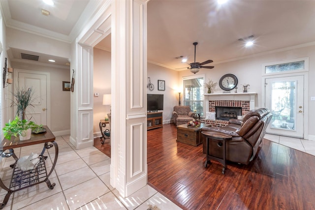 living area featuring a brick fireplace, a ceiling fan, ornate columns, visible vents, and ornamental molding