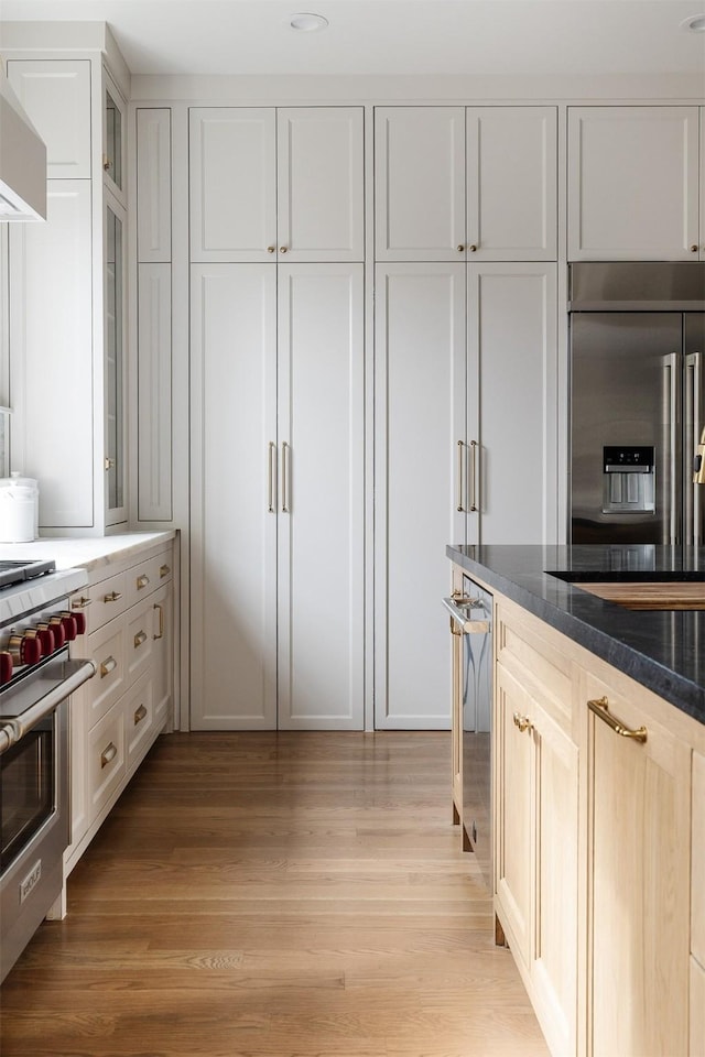 kitchen featuring glass insert cabinets, dark stone countertops, high quality appliances, wall chimney exhaust hood, and light wood-style flooring