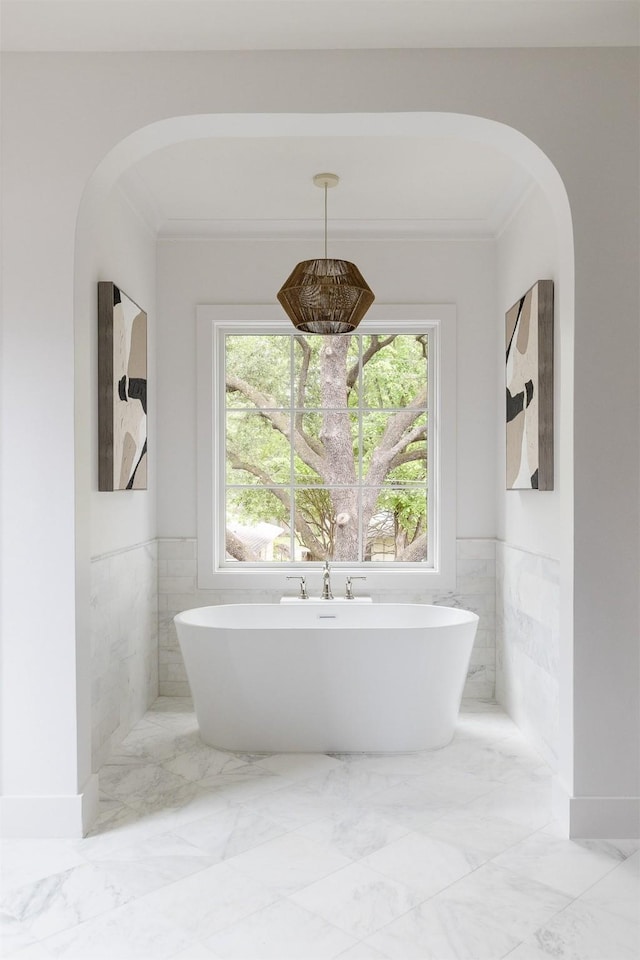 full bath with tile walls, a wainscoted wall, marble finish floor, ornamental molding, and a soaking tub